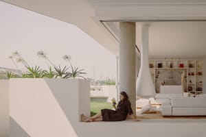 A minimalist living space with concrete walls and large windows, featuring a woman reading a book in a corner.