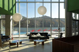 A modern hotel lobby features a glass wall overlooking a serene mountain lake, accompanied by black leather couches and red l