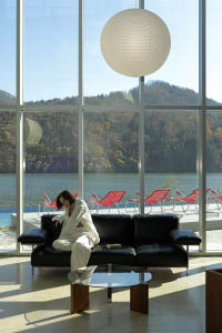 A modern interior featuring a glass window frame, with a black sofa, red lounge chairs, and a large white light fixture.
