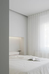 A minimalist bedroom with white walls, curtains, and bedding, featuring a lighted alcove above the bed.