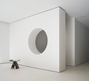 A minimalist white concrete room with a circular cutout in the wall and a wooden stool in a white interior setting.