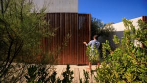 A man standing at the entrance of a modern garden with a wooden fence and green foliage.