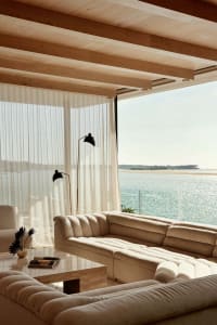 A minimalist living room with a couch, coffee table, floor lamp, and sheer curtains overlooking a body of water.
