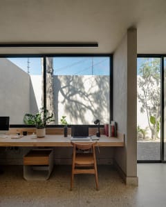 A modern home office featuring a wooden desk with a laptop, lamp, and books, set against a backdrop of large windows showcasi