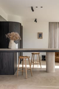 A modern kitchen featuring a concrete countertop, wooden stools, and a vase with dried flowers.