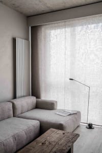 A minimalist living room with a light-colored fabric sofa, concrete radiator, cotton curtains, and a wooden coffee table.