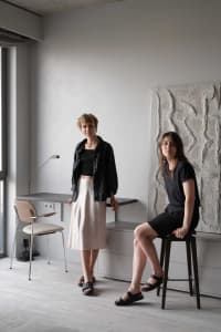 Two women standing and sitting in a minimalist room with white walls, wooden floor, and natural light.