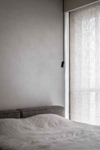 A modern grey bedroom featuring a bed draped in light grey fabric, minimalistic design, and natural light filtering through s