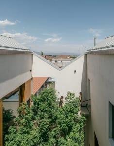 A modern residential house with a concrete exterior, set against a backdrop of greenery and distant mountains.