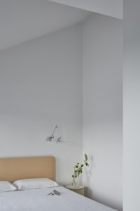 A modern minimalist bedroom featuring a bed with a fabric headboard and white bedding, placed in front of a white wall with a
