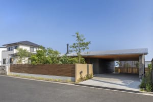 Modern residential house exterior with a concrete front yard surrounded by greenery and a wooden fence.