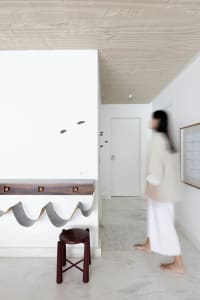 A minimalist interior space with concrete walls and a white ceiling, featuring a wooden stool and a decorative wall piece.