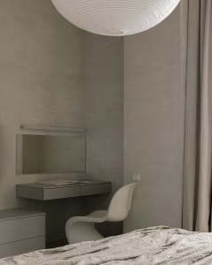 A minimalist bedroom with a bed, chair, desk, and mirror against a textured gray wall.