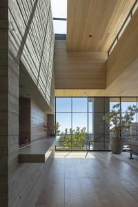 Modern living room with wooden walls, glass windows overlooking greenery, and a concrete fireplace.