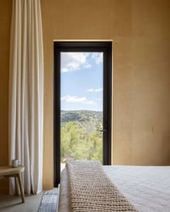 A minimalist bedroom featuring a bed with a brown wooden headboard, a small wooden stool with a mug, and a view of the outdoo
