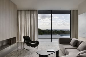 A minimalist living room with a stone floor, a grey sofa, and a black armchair, featuring a large window with view of the sky