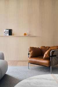 Modern minimalist living room featuring a leather armchair on a textured gray rug with a wooden wall and floating shelf.