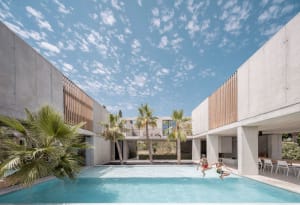 Two people jumping into a swimming pool surrounded by concrete walls with large windows and palm trees.