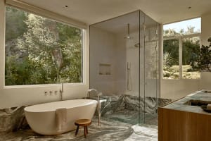 Modern, minimalist bathroom featuring a freestanding marble bathtub and transparent glass shower enclosure, surrounded by nat
