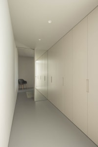A minimalist corridor featuring neutral tone storage units, a wooden floor, and a single armchair in the distance.