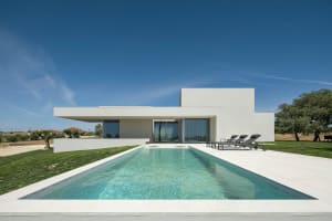 A modern minimalist house with a clean, white exterior featuring a concrete patio and a rectangular pool.