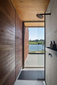 An industrial-style bathroom with a wooden shower area and a view of a tranquil lake through a large glass window.