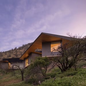 Modern house exterior with wooden roof and glass windows, set against a scenic background during sunset.