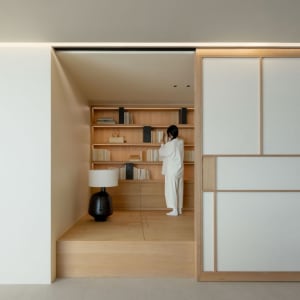 Person wearing white clothes arranging books on wooden shelves in a contemporary library setting.