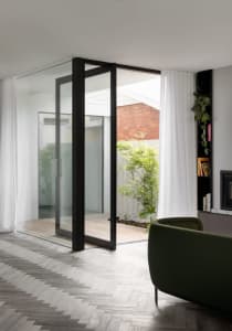 Modern living room with glass and wood elements, featuring a green armchair and white curtains.