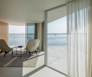 A modern living room with large windows overlooking the sea, featuring a glass and concrete floor, minimalist furniture, and 