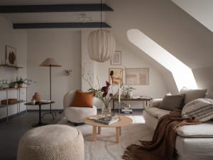 A minimalist living room with a circular wooden coffee table, a white sofa, and a fabric ottoman. The room features a round p