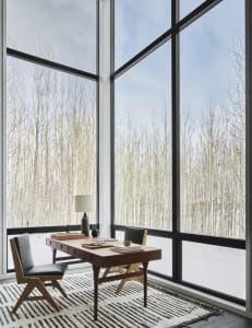 A minimalist home office with a wooden desk and chair, featuring large windows with a snowy forest view.