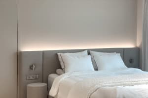 A modern minimalist bedroom featuring a white bed with soft lighting and a light grey upholstered wall