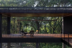 Three people sitting on a wooden deck surrounded by trees under modern industrial architecture.