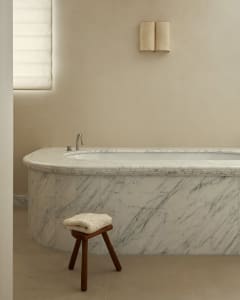 Modern minimalist bathroom featuring a white marble bathtub, a wooden stool with a folded towel, and a sconce on the wall.
