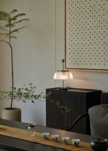 A minimalist living room with wood furniture, a potted plant, a lamp, and a table with cups and a pitcher.