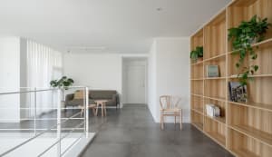 A modern minimalist living room with a grey couch, wooden chair, and bookshelf.