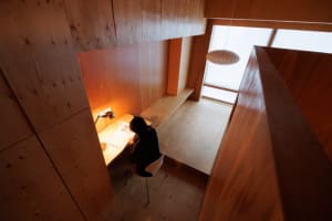 A minimalist workspace with a wooden desk and chair, illuminated by a desk lamp, in a room with wooden panels and a window.