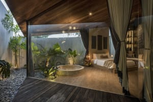 Minimalist bedroom featuring glass walls and a wooden floor, with a circular bathtub in a garden setting.