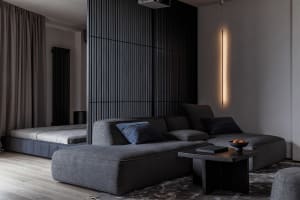 Modern living room with a gray sectional couch, dark wood floor, and a bed behind a slatted privacy screen.