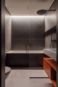 Modern, minimalist bathroom with concrete walls, a floating vanity with orange accents, and a sleek white bathtub.