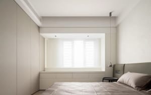 Neutral minimalist bedroom featuring a bed with a simple grey headboard, a window with white shutters, and built-in storage u
