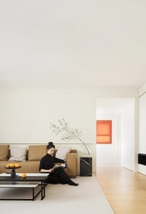 A minimalist living room with neutral tones, featuring a person sitting on a brown sofa, reading a magazine, and placing oran