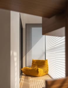 Modern living room featuring a yellow upholstered chair on a wooden floor with sunlight streaming through horizontal blinds.