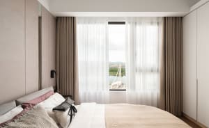 A serene bedroom featuring modern design with light-colored fabric and a glass window overlooking a scenic view.