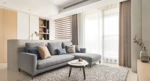 A modern minimalist living room with a gray sectional sofa, beige cushions, and a round coffee table on a patterned rug.