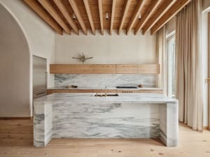Modern residential kitchen with a large white marble island, wooden ceiling beams, and light wooden floors.
