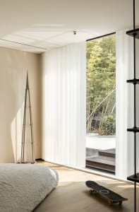 A minimalist bedroom with white curtains and a view of nature, featuring wood flooring and a skateboard.