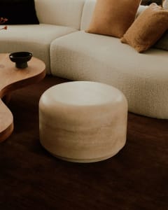 A modern living room with a white sectional sofa, brown throw pillows, a wooden curved coffee table, and a concrete cylindric