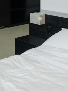 Modern minimalist bedroom featuring a textured black wooden platform bed with white bedding and a geometric lamp on the headb